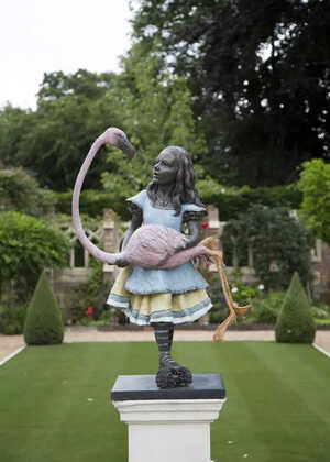 Bronze sculpture of Alice in a blue dress holding a flamingo in a garden.
