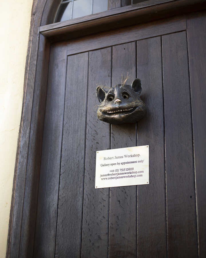 The front door of the Robert James Workshop Gallery with the Cheshire Cat Face Sculpture attached and an appointment-only sign below