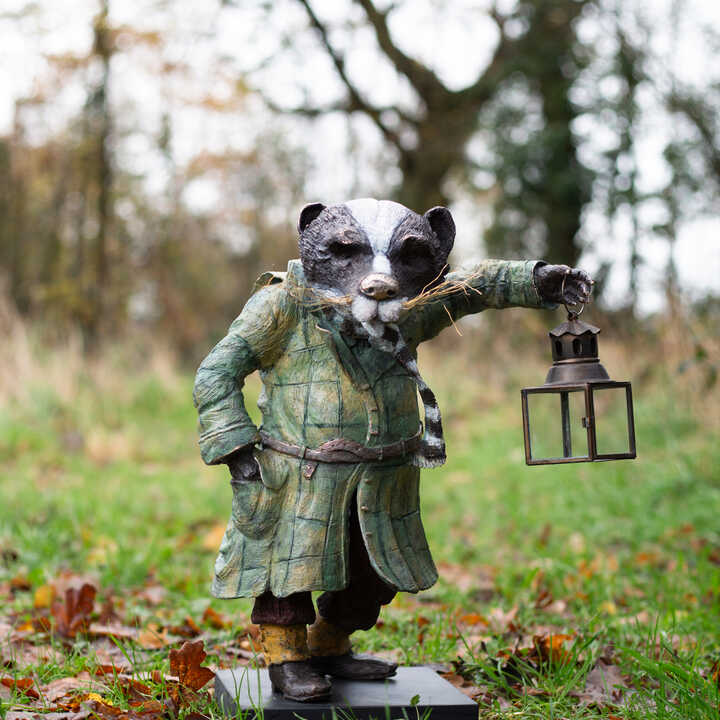 Mr. Badger sculpture in a coat holding a lantern, standing on grass among fallen leaves.