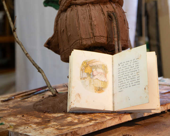 Open book with illustrations of Mrs Tiggy-Winkle on a wooden table, next to a sculpted figure.