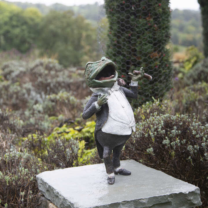 Mr. Toad sculpture wearing a tuxedo, standing on a stone pedestal in a garden.