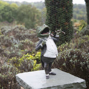 Mr. Toad sculpture wearing a tuxedo, standing on a stone pedestal in a garden.